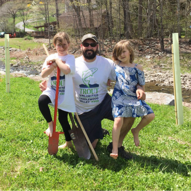 A smiling, bearded man poses with his two daughters near a stream.
