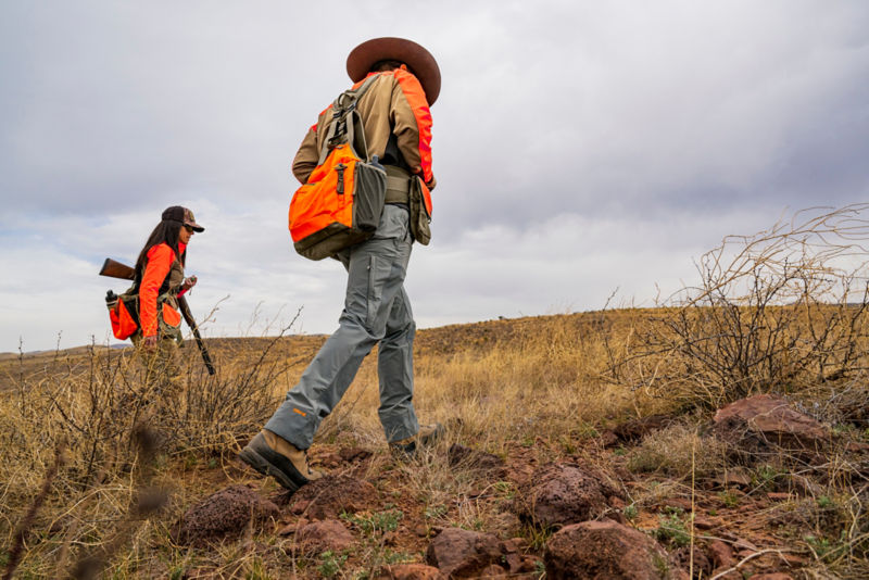 A man out in the field carrying a gun in full hunting apparel.