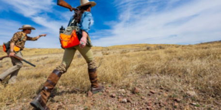 A man and a woman wearing hunting gear walking through a field of dry grass.