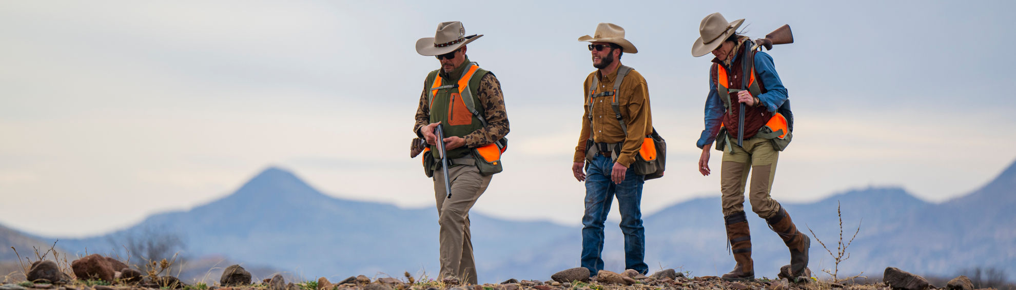 Three hunters wearing hunting gear walking in an open fi.eld with mountains in the background.