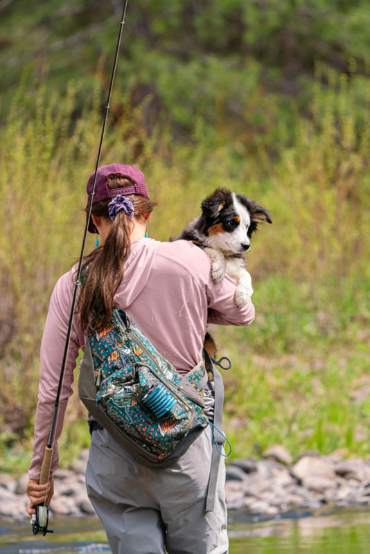A woman holding a dog wearing a sling pack headed out fishing