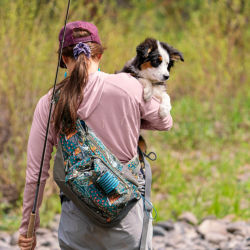 A woman holding a dog wearing a sling pack headed out fishing