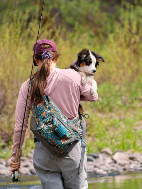Women walking in a river holding a puppy and fly rod and wearing an Orvis sling pack.