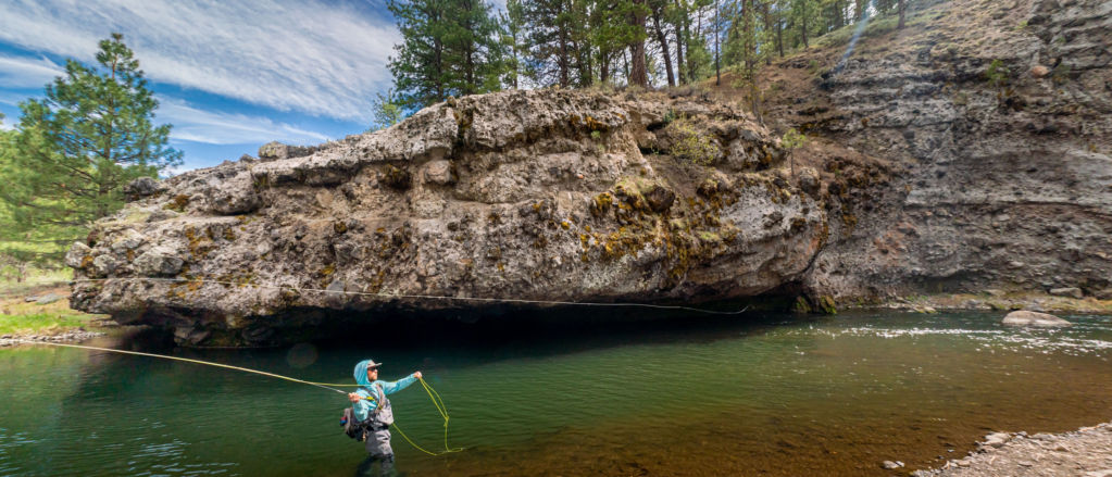 An angler in waders and a hoodie fishes a nook beneath a rock outcropping.