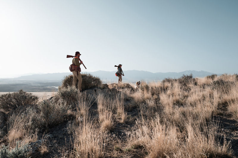 Two hunters stand on a rise overlooking fields surrounded by mountains.