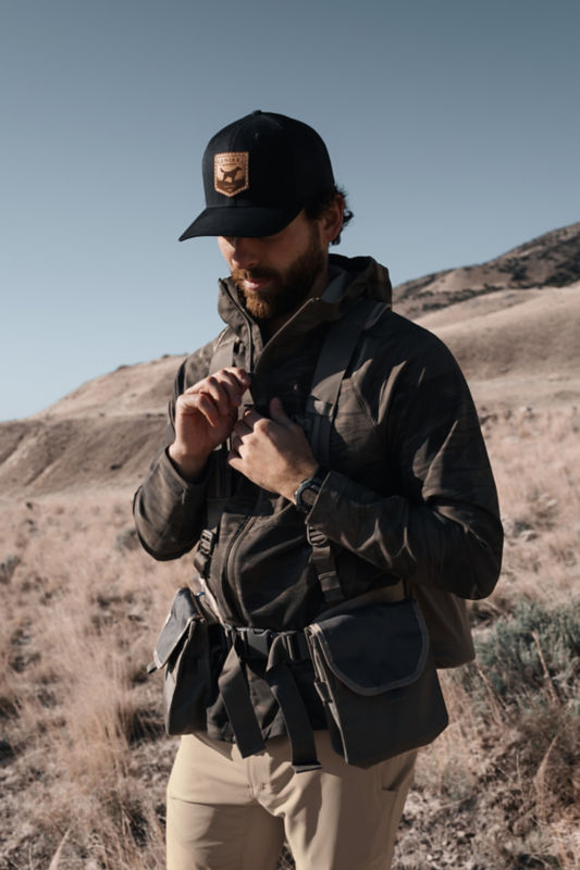 A hunter zipping up his hunting vest as he's standing on a hill.