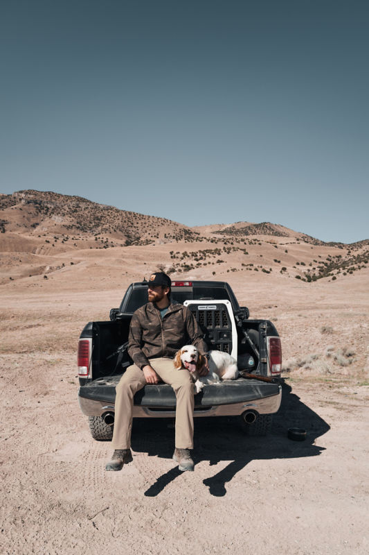 A man wearing a camo jacket and tan pants is sitting with his dog on the tailgate of a truck.