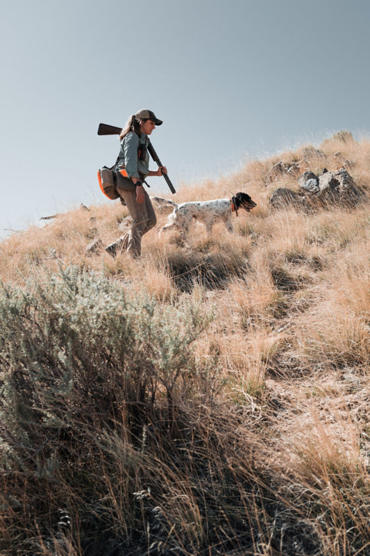 A hunter climbs a hill with her shotgun over her shoulder and her dog by her side.