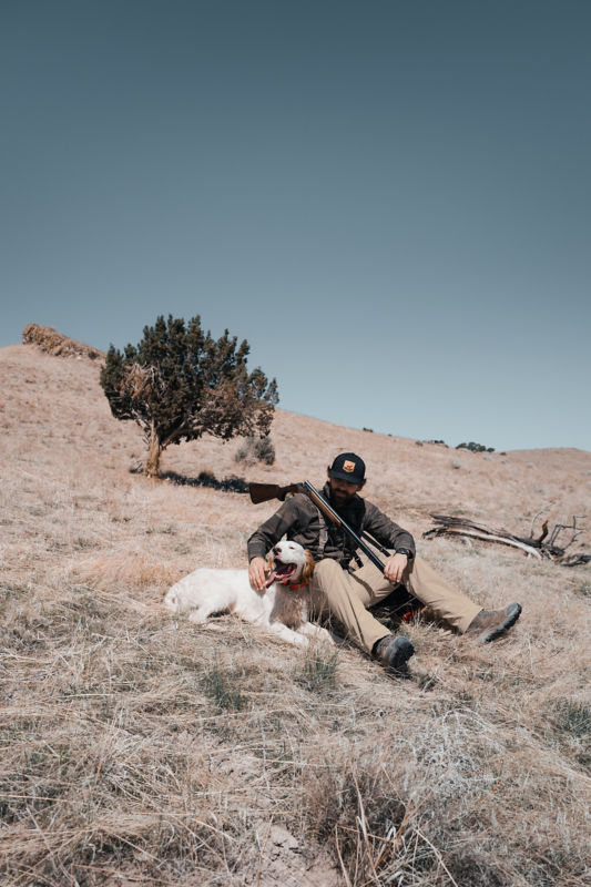 A hunter rests on the ground with his arm around his happy dog.