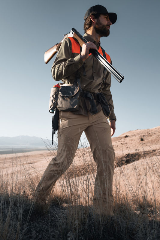 A hunter walking in a grassy field holding a shotgun over his shoulder.
