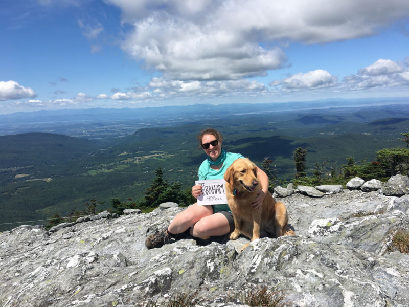 A woman and her golden sitting at the top of a mountain surrounded by blue skies.