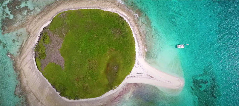 A drone-view of a small round green island with a boat anchored in turquoise waters.