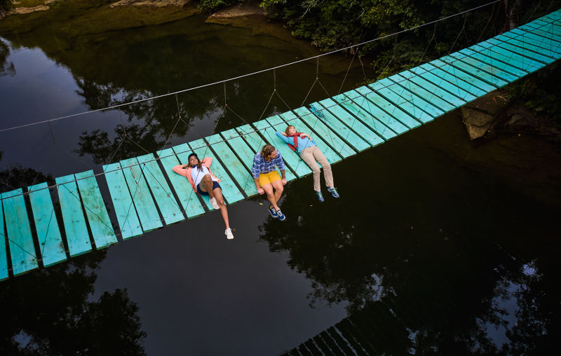 Three hikers pause for a rest on a turquoise bridge over a river.