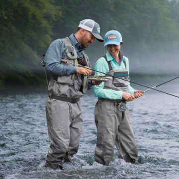 Two anglers pick through a fly box as they wade through a river.
