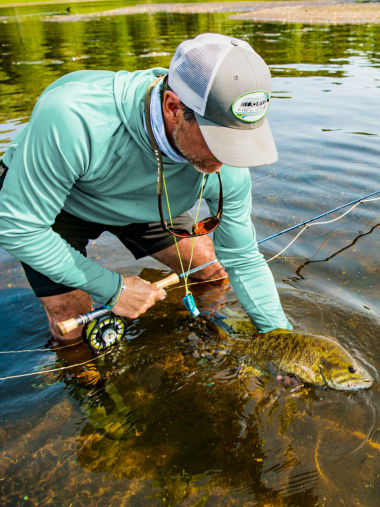 A wet-wading angler holds their catch along the water's surface with one hand and their fly rod in the other hand.