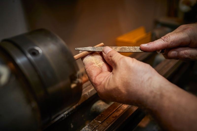 Hands filing a wooden dowel at a workbench.