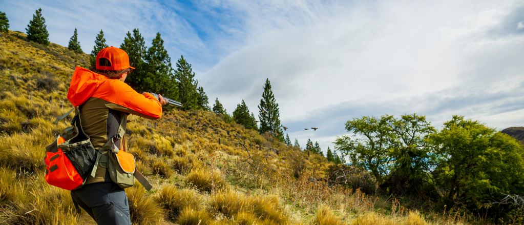 An angler stands on a hillside field aiming her shotgun