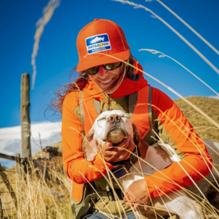 A woman wearing blaze orange crouches in a field to hug her white dog.