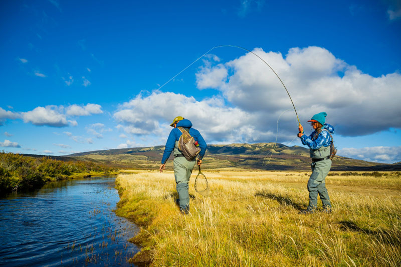 Two people fishing from the bank of a river in an open field 