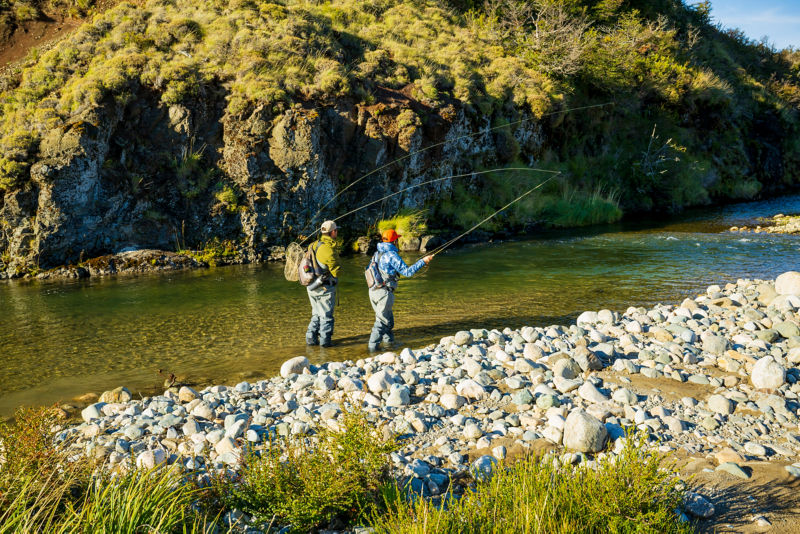 Two anglers fish from ankle-deep in a shallow green river with a gravel beach.