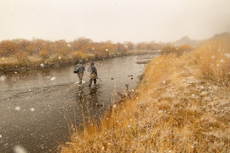 Two anglers wade through a river as it snows.