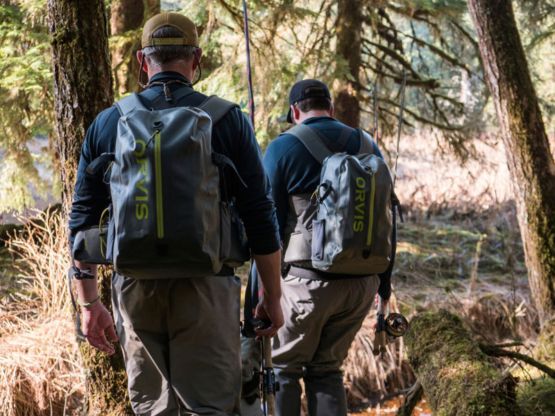 Two people walking along a river wearing Orvis packs and Orvis waders.