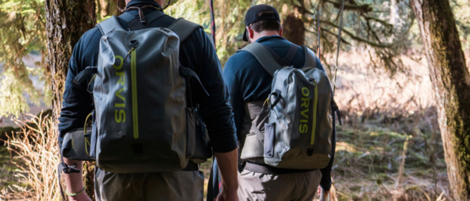 Two people walking along a river wearing Orvis packs and Orvis waders.