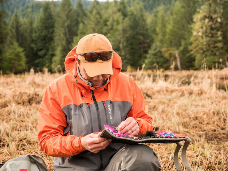 An angler sits on dried grass picking through a fly box for the perfect fly.