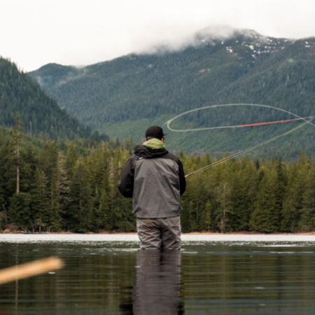 An angler casting a fly rod knee-deep in a lake