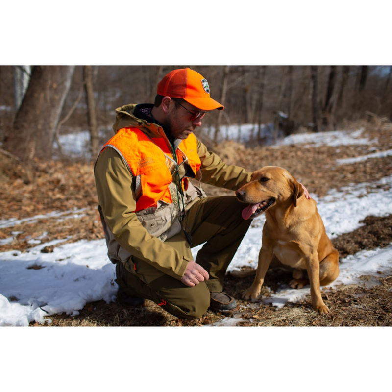 A man in hunting gear crouching down on the snowy ground petting a yellow dog