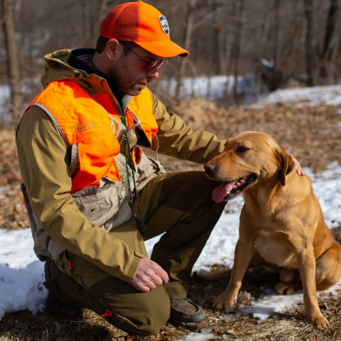 A man in hunting gear crouching down on the snowy ground petting a yellow dog