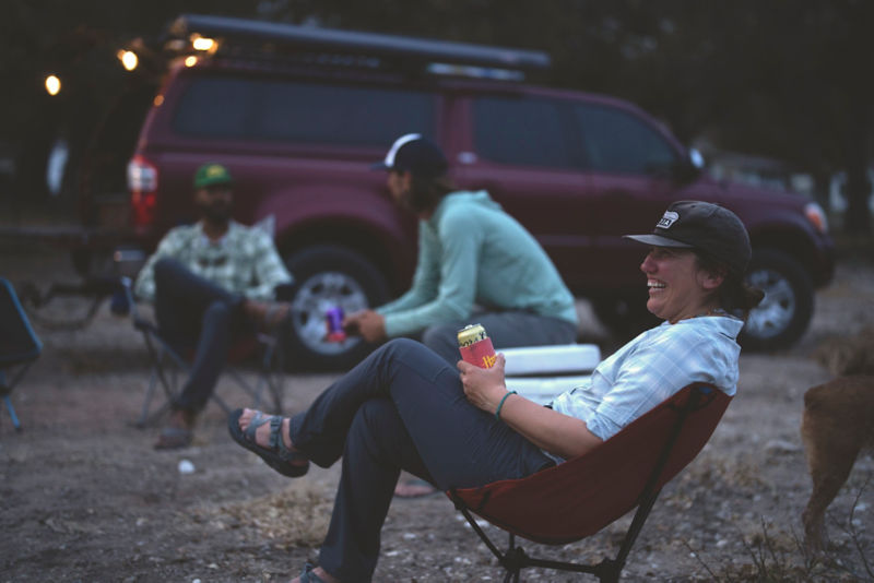 A woman lounging with a beer in a camp chair chatting with a group next to a truck at dusk