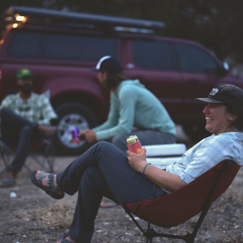 A woman lounging with a beer in a camp chair chatting with a group next to a truck at dusk