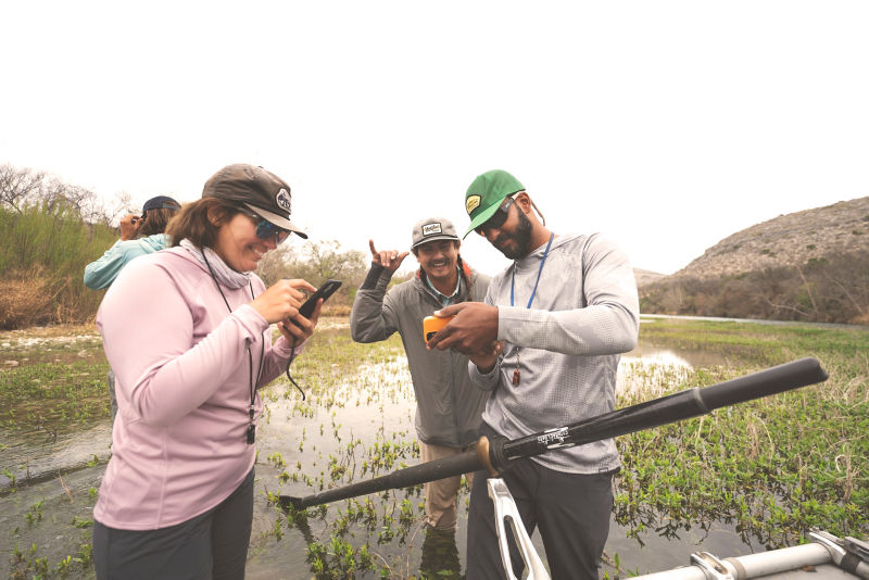 Three people stand next to a row boat sharing photos from their boat trip