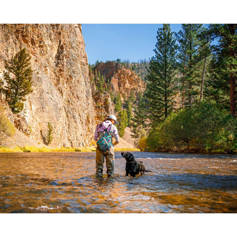 A woman in waders casts her fly rod in the water along with her black lab.