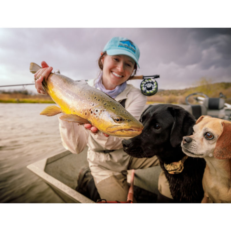 A woman holding up a large spotted fish while two dogs look beyond.
