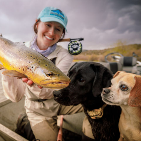 A woman holding up a large spotted fish while two dogs look beyond