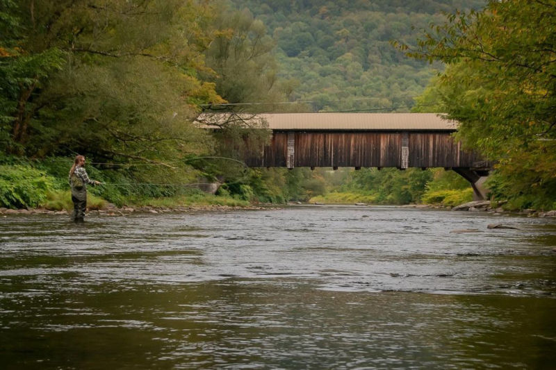 An angler fly fishing in a river by a brown covered bridge.