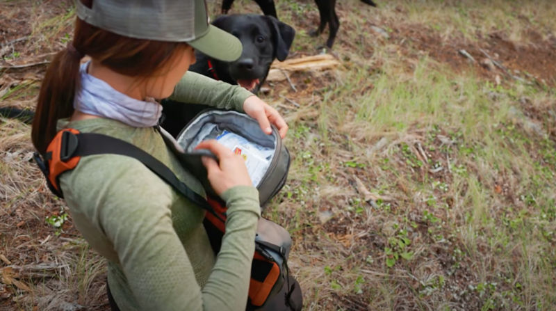 A woman standing out in a field near her black dog wearing a day pack with first aid items