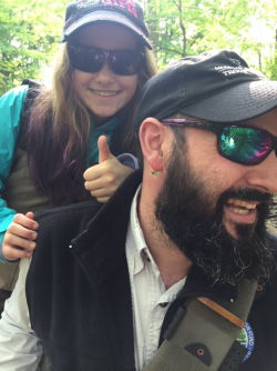 A bearded man wearing sunglasses smiles as his daughter gives the thumb's up, behind him.