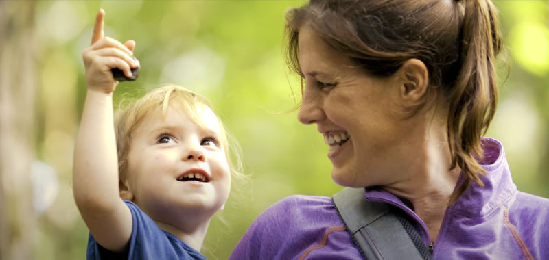 A smiling mother holds a small child who is pointing upwards.