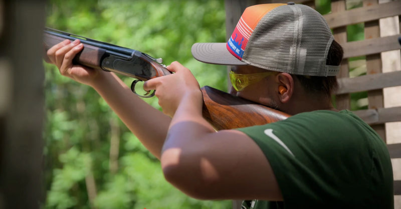 A man in a ball cap sighting a shotgun at a range