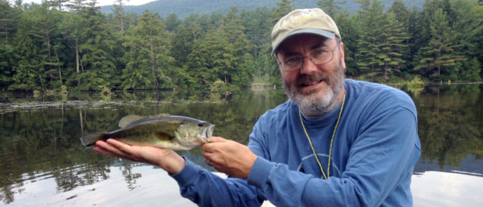 Phil Monahan holding a small bass while sitting in a canoe on a pond