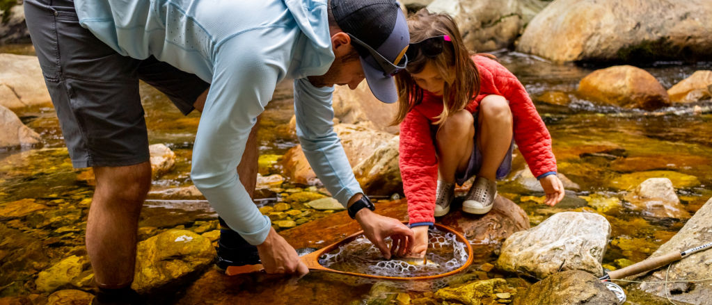 A father and daughter standing in a creek crouched down inspecting a fish in a fishing net