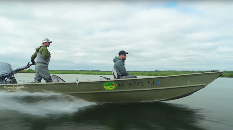 Two anglers wearing waders in a speeding boat under a cloudy sky.