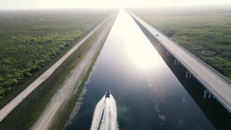 A boat follows a straight shot of river between two roads