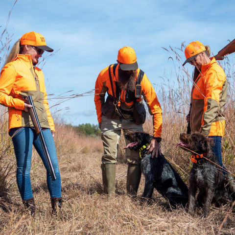 Three people in hunting gear standing in a field, one person petting a black dog